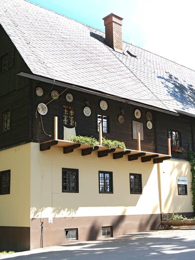 Ein traditionelles, zweigeschossiges Gebäude mit einem schönen, schattigen Außenbereich. Die Fassade ist in zwei Farben gestrichen und wird von Bäumen umgeben. | © TV Region Graz - Donner