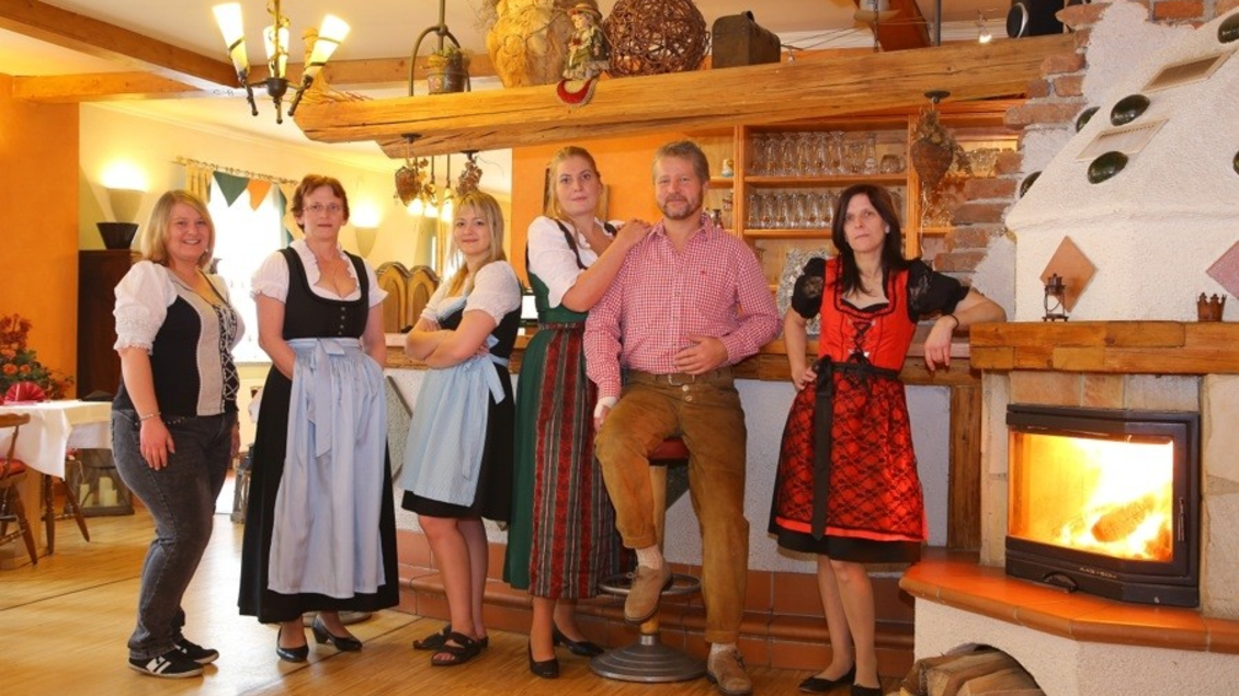 Eine Gruppe von sechs Personen in traditionellen Trachten steht in einem gemütlichen Restaurant. Im Hintergrund ist ein Kamin und eine rustikale Einrichtung zu sehen. | © Gasthaus Ruckenstuhl