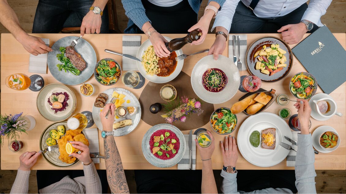 Ein Tisch voller köstlicher Gerichte, umgeben von Menschen, die gemeinsam essen. Die Speisen sind bunt und vielfältig, was eine einladende Atmosphäre schafft.