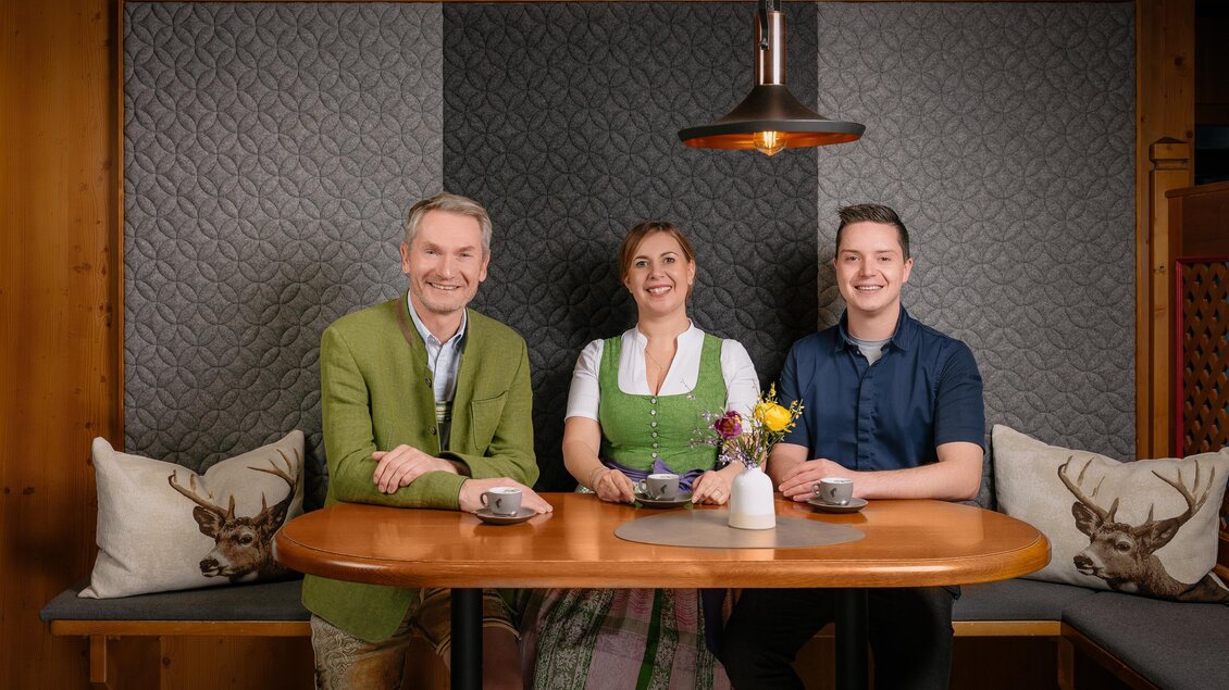 Drei Personen sitzen an einem Tisch in einem gemütlichen Raum. Sie tragen traditionelle Kleidung und wirken freundlich und aufgeschlossen.