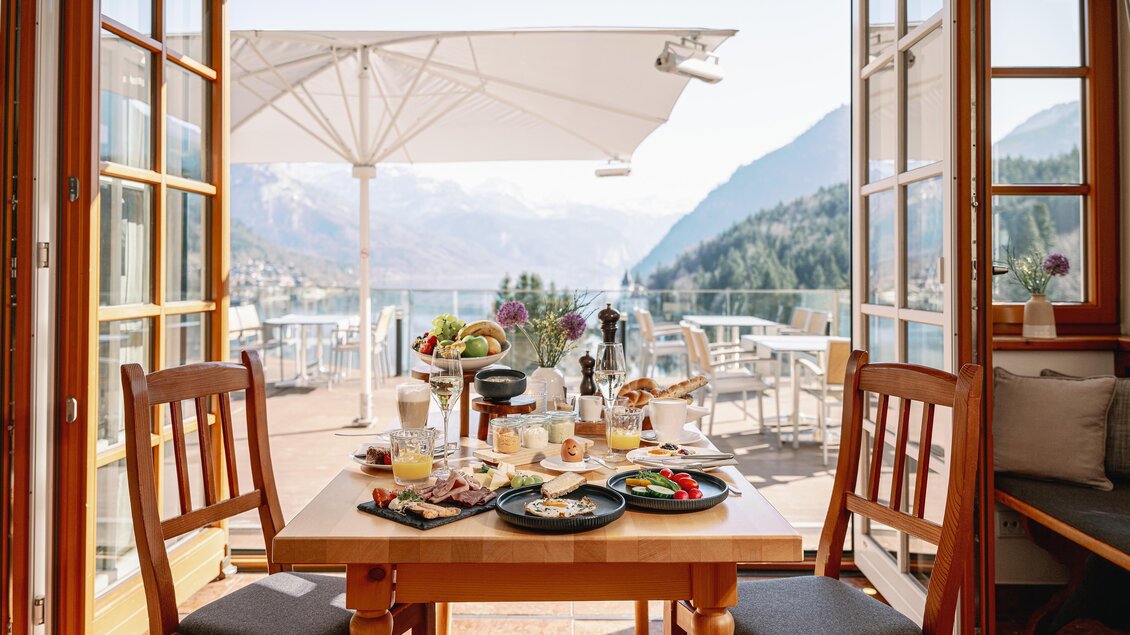 Ein gemütlicher Tisch mit Frühstücksangebot in einem offenen Raum. Im Hintergrund sind Berge und ein klarer Himmel sichtbar.