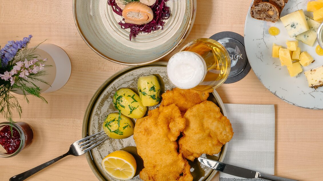 Ein Teller mit knusprigem Schnitzel, Kartoffeln und Zitronenscheiben. Dazu ein Glas Bier und verschiedene andere Speisen auf dem Tisch.