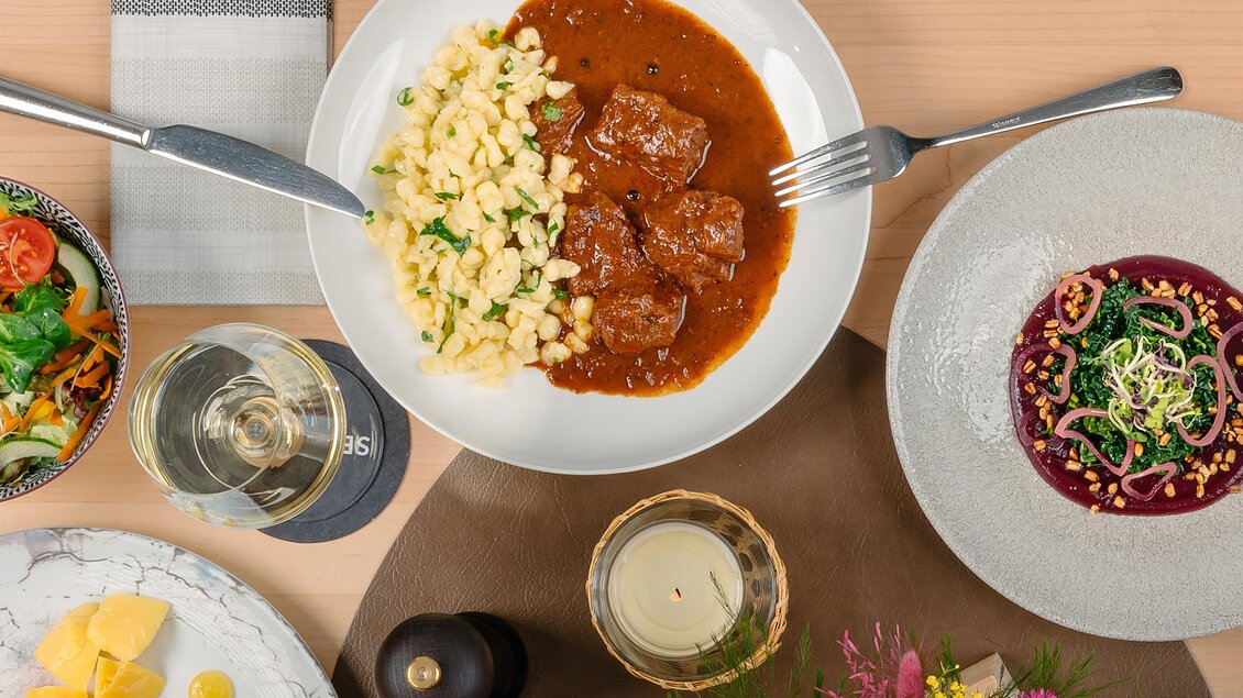 Ein Teller mit Fleischgericht in Sauce, serviert mit Spätzle. Daneben sind Salat, Wein und Desserts auf dem Tisch.