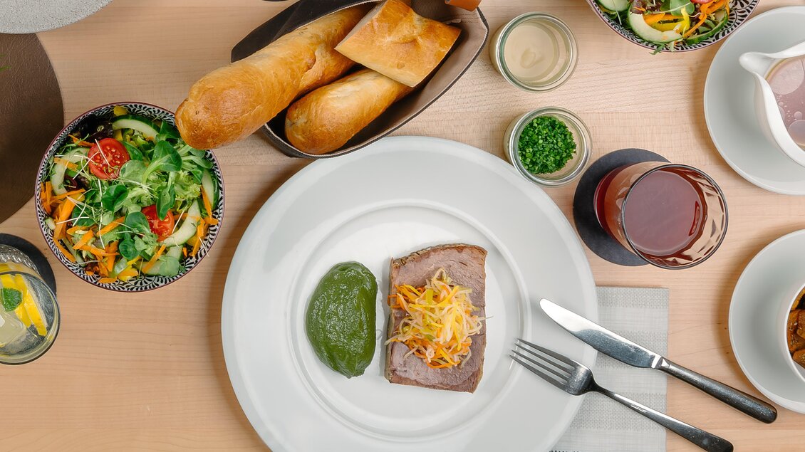 Ein stilvolles Tischarrangement mit einer Hauptspeise aus Fleisch und grüner Soße. Dazu gibt es frische Salate, Brötchen und Getränke.