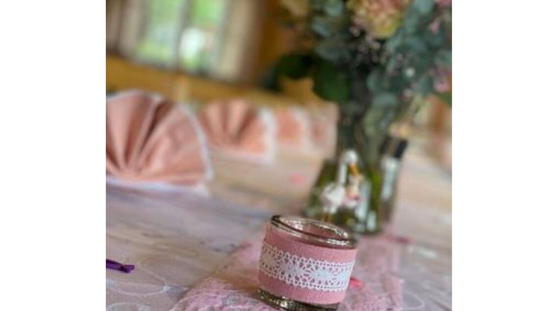Eine schön dekorierte Tischgedeck mit einem rosa Teelicht und einer Blumenvase im Hintergrund. Der Tisch ist mit einem weißen Tischtuch und Faltservietten geschmückt. | ©  Schneiderwirt_Kainzinger