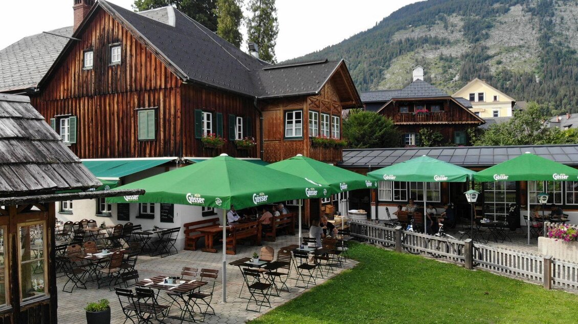 Ein charmantes Gasthaus mit Holzfassade und grünen Sonnenschirmen im Garten. Die Umgebung ist von Bergen und Bäumen umgeben. | ©  Schneiderwirt_Kainzinger