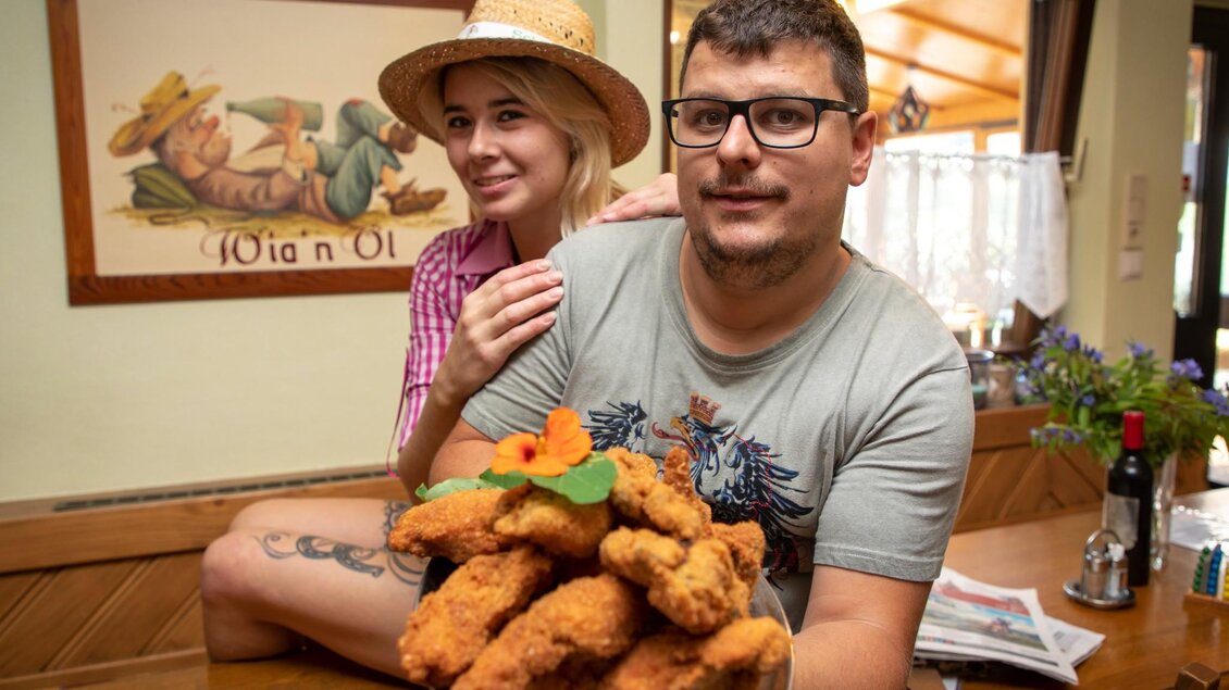 Ein fröhliches Paar posiert in einem Restaurant mit einem witzigen Wandgemälde im Hintergrund. Die Frau trägt einen Strohhut und hat eine fröhliche Ausstrahlung. | © TV Region Graz - Lunghammer