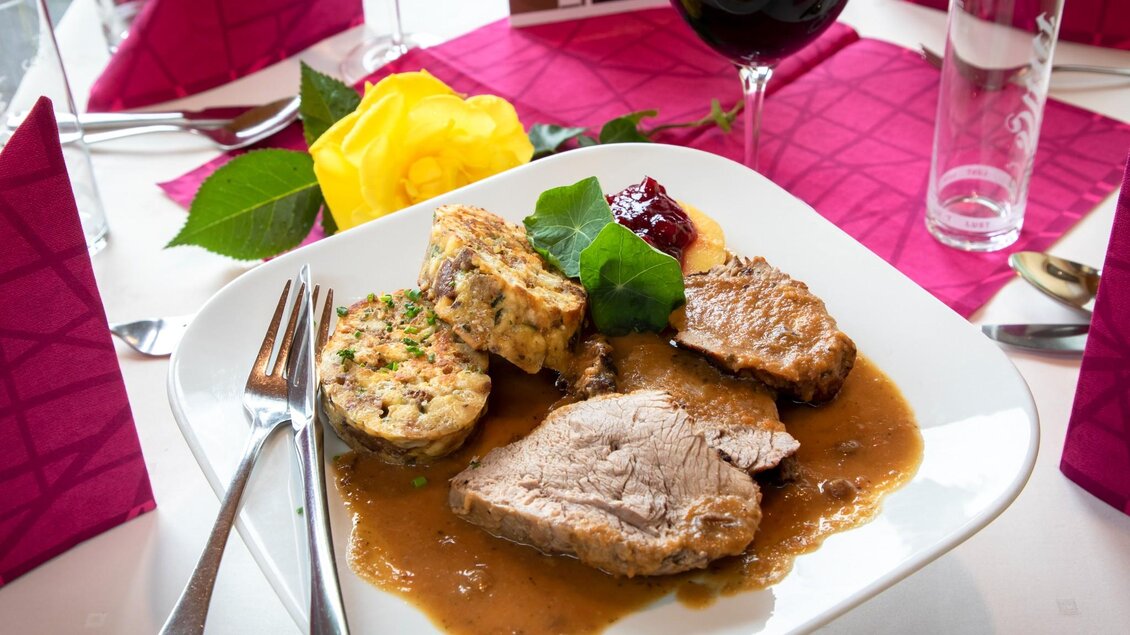 Ein Teller mit saftigem Fleisch in Sauce, serviert mit Knödeln und Preiselbeeren. Im Hintergrund sind bunte Bilder von Sehenswürdigkeiten zu sehen. | © TV Region Graz - Lunghammer
