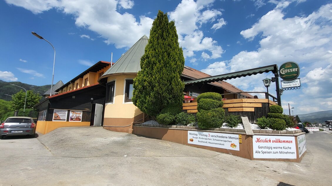 Ein Restaurantgebäude mit einem begrünten Außenbereich und einem Parkplatz. Der Himmel ist blau mit einigen Wolken. | © TV Hochsteiermark