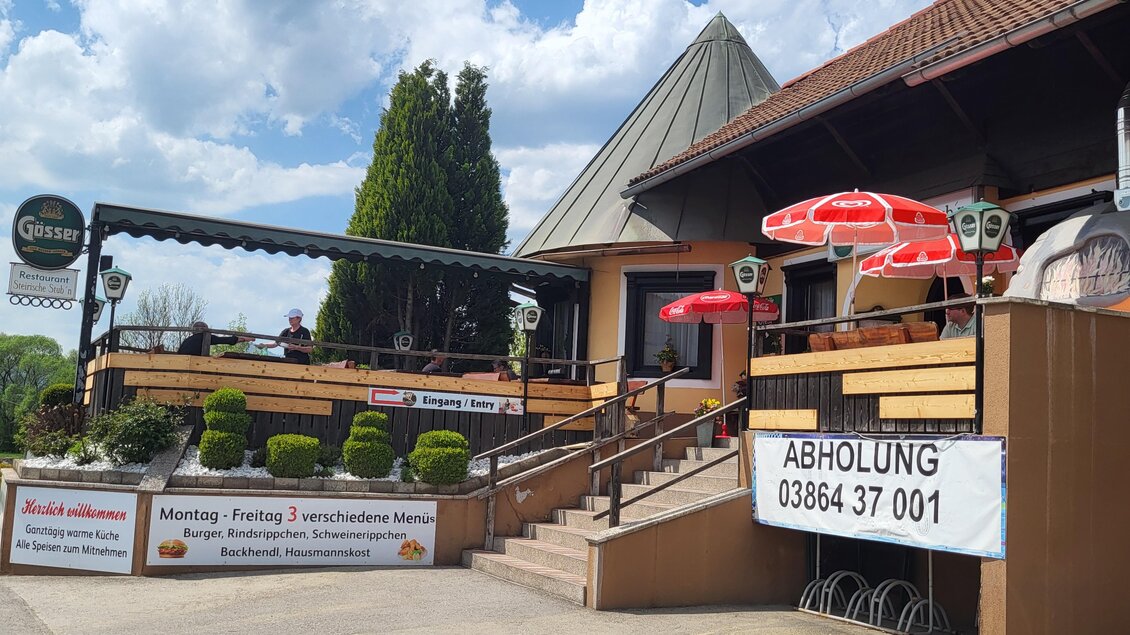 Ein Restaurant mit einer einladenden Terrasse und roten Sonnenschirmen. Im Hintergrund sind einige grüne Pflanzen und ein blauer Himmel mit Wolken sichtbar. | © TV Hochsteiermark