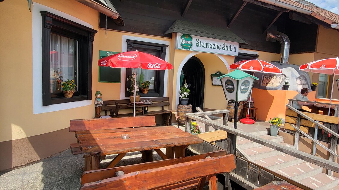 Ein gemütlicher Außenbereich eines Restaurants mit Holztischen und roten Sonnenschirmen. Die Fassade des Lokals ist hell und einladend, umgeben von blauen Himmel und wenigen Wolken. | © TV Hochsteiermark