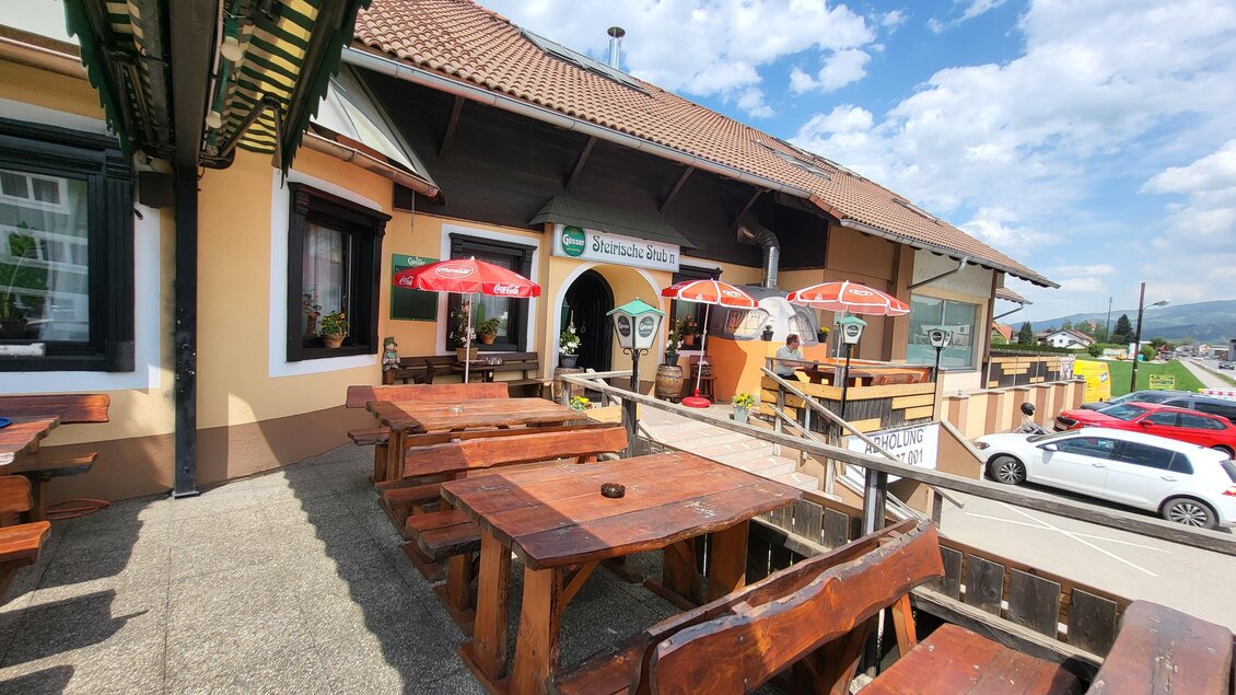 Ein gemütliches Restaurant mit Holzterrasse und roten Sonnenschirmen. Im Hintergrund ist ein blauer Himmel mit einigen Wolken zu sehen. | © TV Hochsteiermark