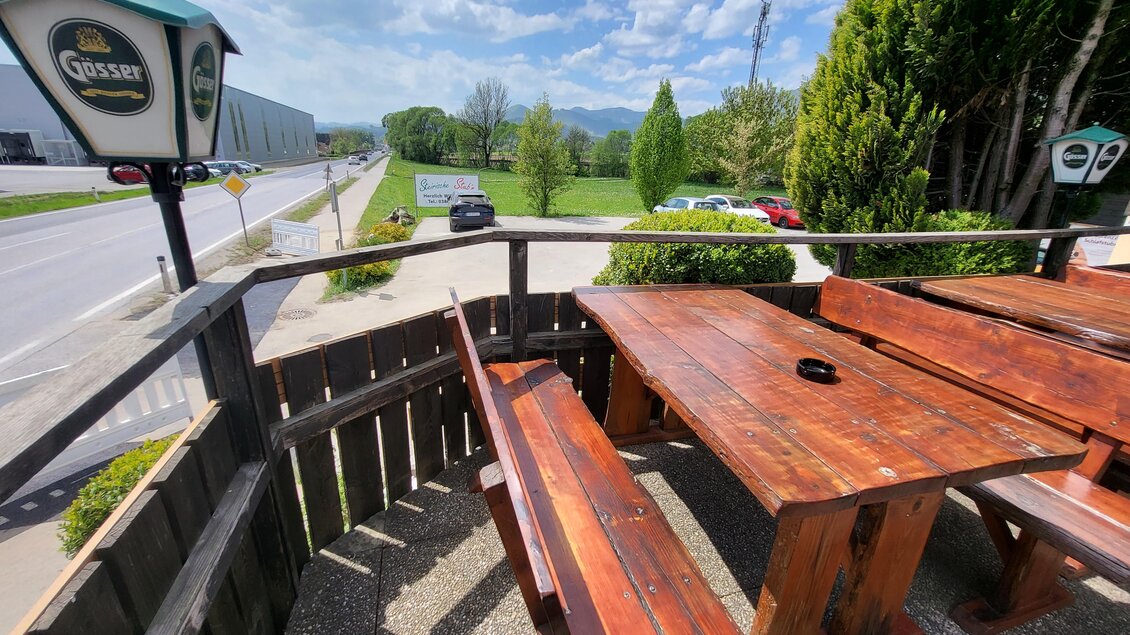 Ein gemütlicher Außenbereich mit einer Holztischgruppe. Im Hintergrund sind grüne Bäume und ein blauer Himmel sichtbar. | © TV Hochsteiermark