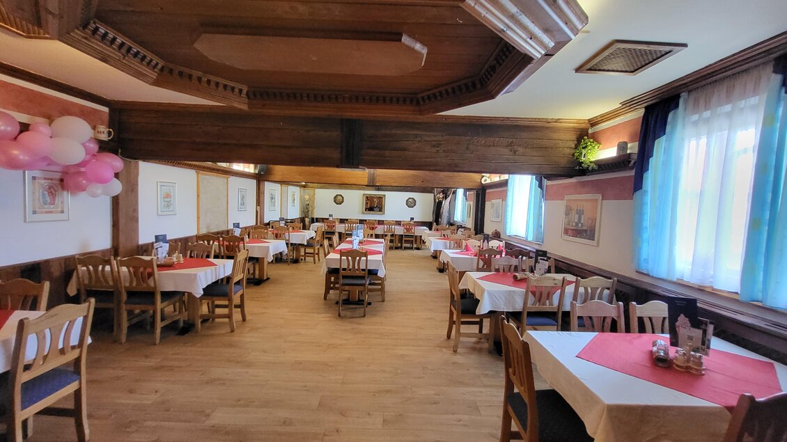 Ein gemütliches Restaurant mit Holzdekor und Tischen, die mit weißen und roten Tischdecken gedeckt sind. Rosa Dekorationen schmücken den Raum und große Fenster sorgen für viel Tageslicht. | © TV Hochsteiermark