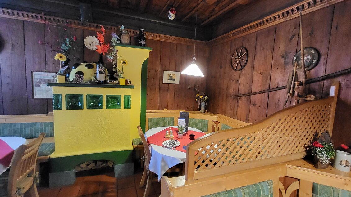Ein gemütliches Restaurant mit Holzverkleidung und traditionellem Dekor. Es gibt eine grüne Ofenbank und Tische mit Tischdecken. | © TV Hochsteiermark