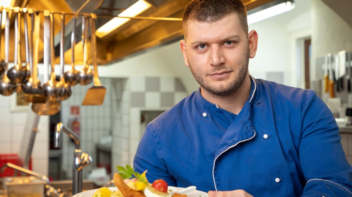 Ein Koch steht in einer modernen Küche und lächelt stolz. Er hält einen Teller mit frischen Zutaten in der Hand. | © TV Region OberGraz - Lunghammer