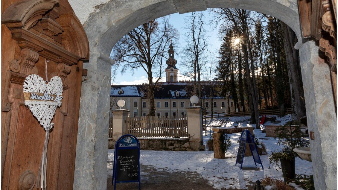 Ein malerischer Blick auf ein historisches Gebäude durch ein Tor, umgeben von schneebedeckten Bäumen. Die Sonne scheint durch die Bäume und verleiht der Szene eine warme Atmosphäre. | © TV Region OberGraz - Lunghammer