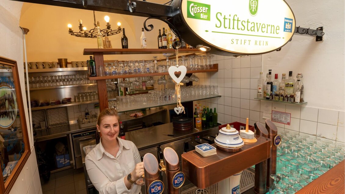 Eine freundliche Kellnerin steht hinter einer Bar. Der Raum ist gut beleuchtet und enthält eine Auswahl an Getränken und Gläsern. | © TV Region OberGraz - Lunghammer