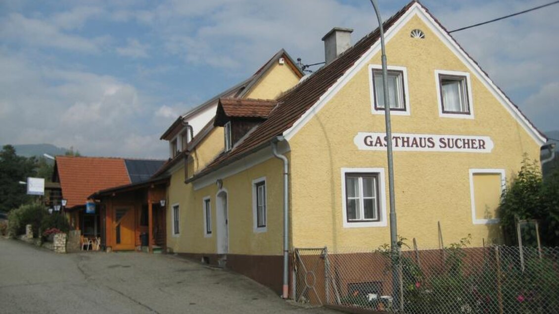 Ein gelbes Gasthaus mit einem einladenden Eingangsbereich. Die Straße ist ruhig und gepflastert, umgeben von einer grünen Landschaft. | © Erlebnisregion Murtal