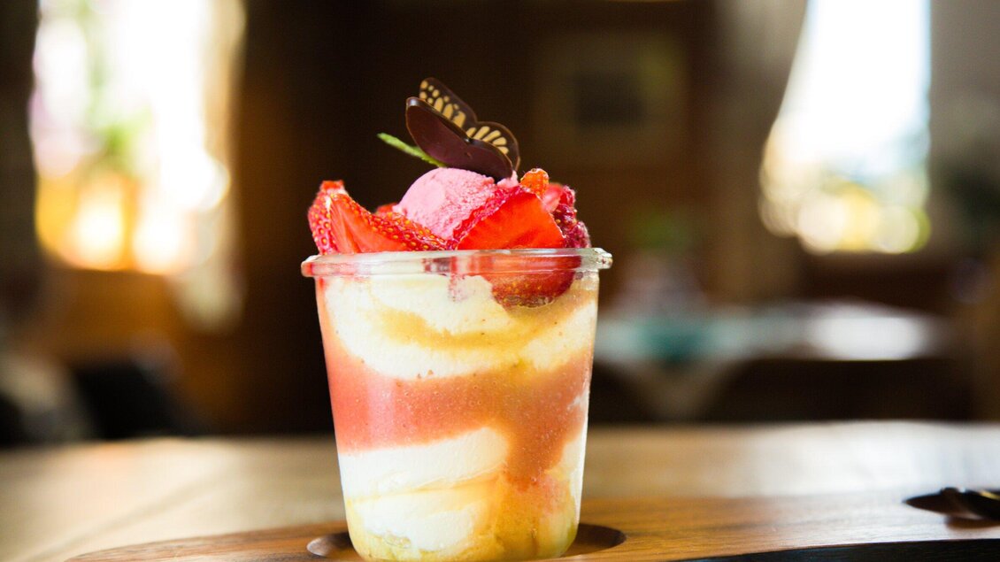 Ein attraktives Dessert in einem Glas mit Schichten aus Eiscreme und frischen Erdbeeren. Oben ist eine essbare Blüte und ein Schokoladenstückchen dekoriert. | © Geopho