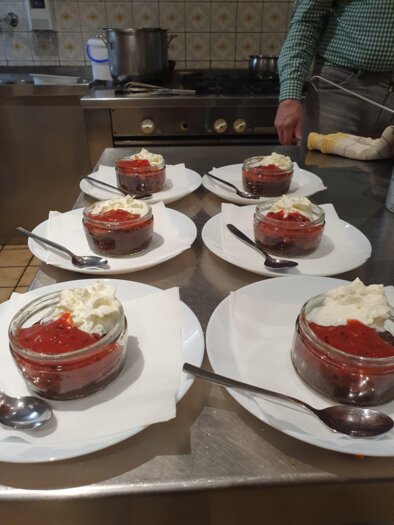 Sechs Gläser mit Dessert auf weißen Tellern, garniert mit Sahne und fruchtiger Soße. Im Hintergrund sind Kochutensilien sichtbar. | © Emil