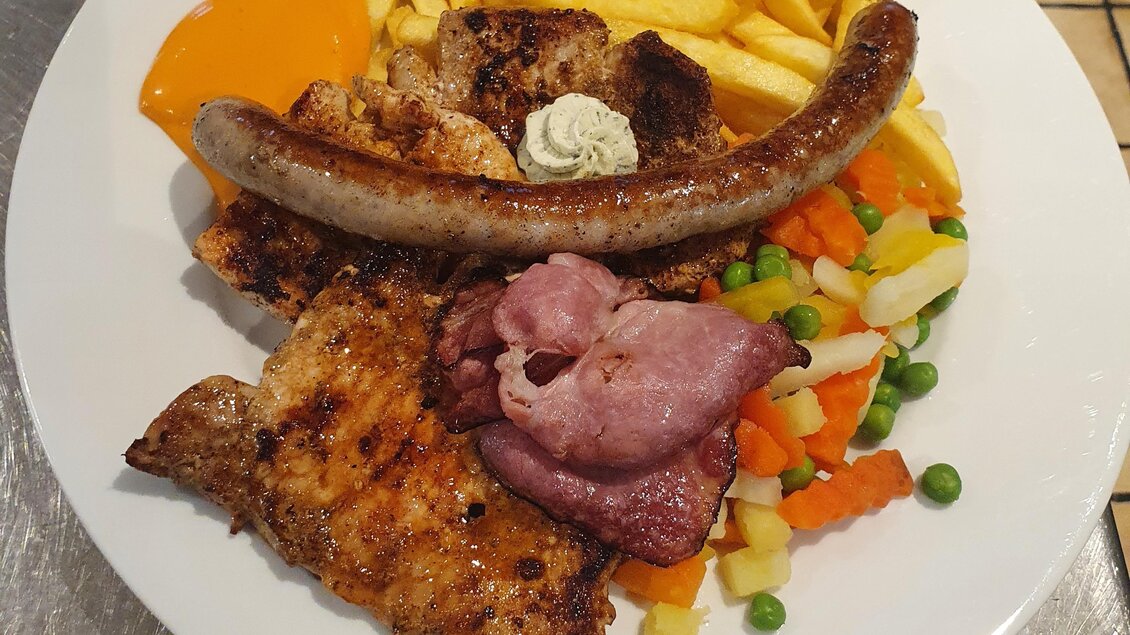 Ein Teller mit gebratenem Fleisch, einer Wurst, Pommes frites und buntem Gemüse. Dazu gibt es eine Portion Sauce. | © Emil