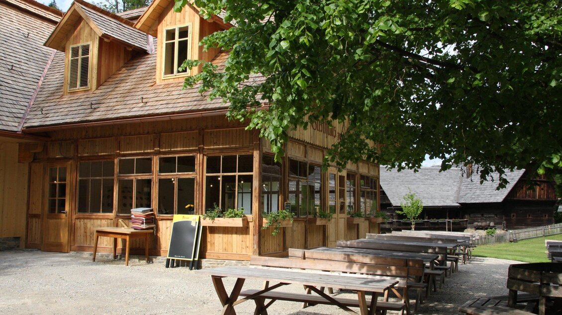 Ein gemütliches Holzhaus mit großen Fenstern und einem begrünten Außenbereich. Mehrere Holztische stehen im Freien, umgeben von Bäumen. | © OeFM Stuebing