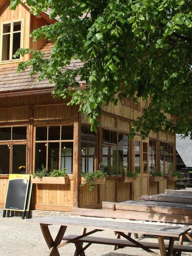Ein gemütliches Holzhaus mit großen Fenstern und einem begrünten Außenbereich. Mehrere Holztische stehen im Freien, umgeben von Bäumen. | © OeFM Stuebing