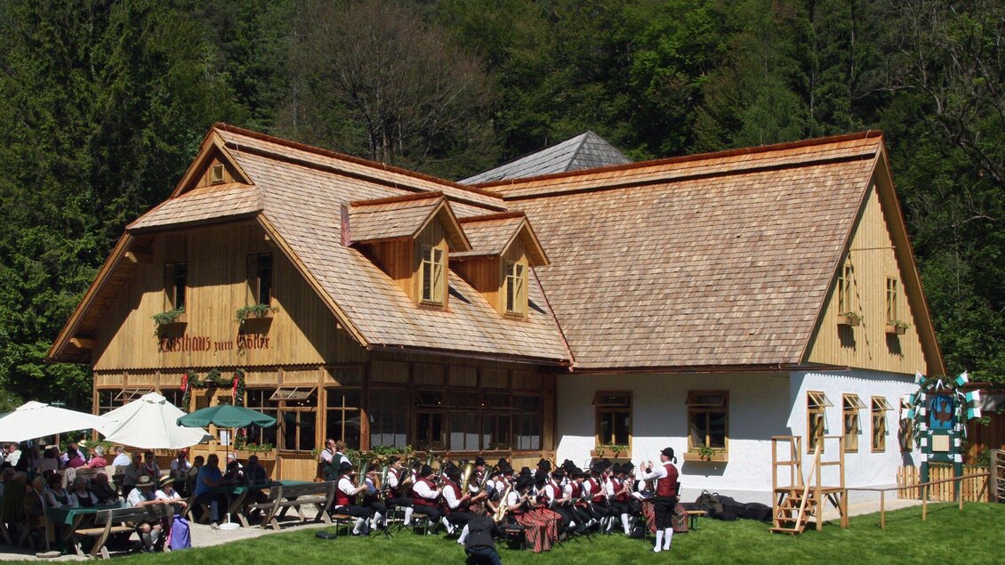 Ein traditionelles Holzhaus mit einem schönen Dach in der Natur. Vor dem Haus sitzt eine Gruppe von Menschen in Trachten und genießt die Atmosphäre. | © OeFM Stuebing