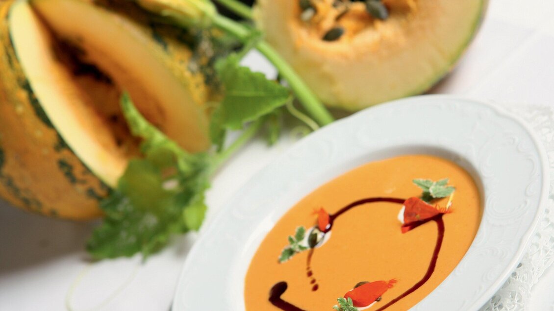 Ein Teller mit cremiger Kürbissuppe, garniert mit Kräutern und einer Sauce. Im Hintergrund liegt ein halbierter Kürbis. | © Graz Tourismus - Harry Schiffer