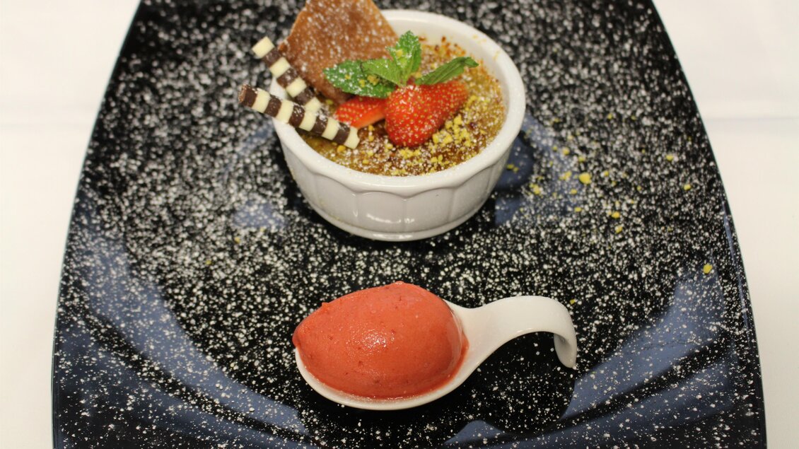 Ein elegantes Dessert auf einem schwarzen Teller, bestehend aus einer Crème brûlée mit frischen Erdbeeren und Dekoration. Nebenan befindet sich eine Portion Erdbeersorbet. | © Gasthaus zur Alten Press