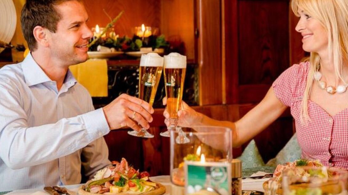 Ein Paar prostet sich mit Gläsern Bier zu, während sie an einem Tisch mit köstlichem Essen sitzen. Im Hintergrund sind gemütliche Restaurantdetails sichtbar. | © Gasthof Bräuer
