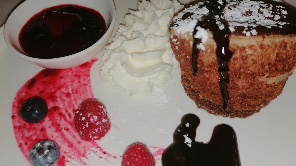 Ein köstliches Dessert auf einem weißen Teller mit einem Schokoladenkuchen, Sahne, frischen Beeren und Fruchtsoße. Der Teller ist dekoriert mit roten und blauen Früchten sowie einer kleinen Schale mit Sauce. | © Gasthof  Bräuer