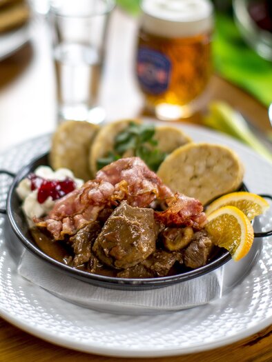 A delicious dish on a white plate, consisting of tender meat, side dishes, and fresh lemon slices. In the background, drinks and other foods are visible. | © Tourismusverband Murau