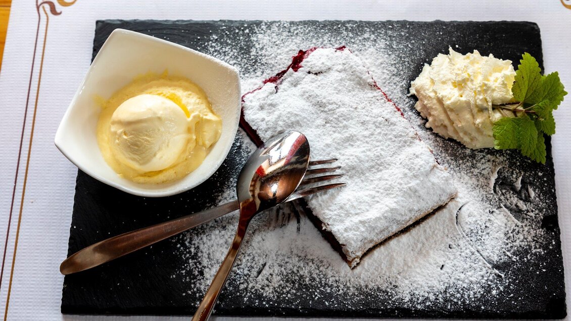 Ein köstliches Stück Kuchen mit Puderzucker, serviert auf einem schwarzen Teller. Dazu gibt es eine Kugel Vanilleeis und einen kleinen Klecks Schlagsahne. | © TV Region Graz - Lunghammer