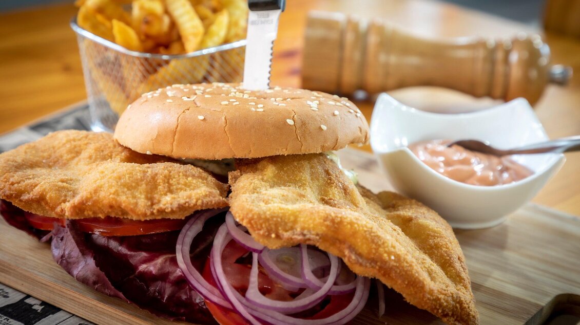 Ein saftiger Burger mit paniertem Fleisch, Tomaten und Zwiebeln, serviert auf einem Holzbrett. Dazu gibt es Pommes frites und eine Schale mit Sauce. | © TV Region Graz - Lunghammer