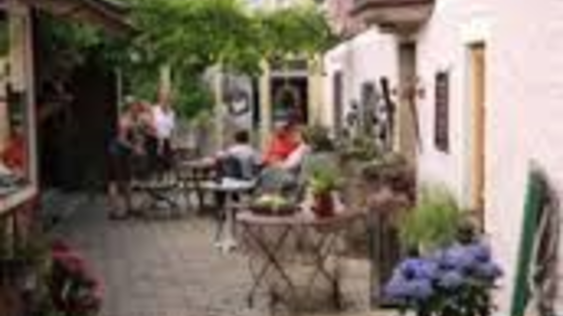 Eine malerische Gasse in einem Dorf mit traditionellen Gebäuden. An den Seiten stehen Tische und Blumen, während Menschen gemütlich sitzen und die Umgebung genießen. | © Gasthof Angermaier - Winklwirt
