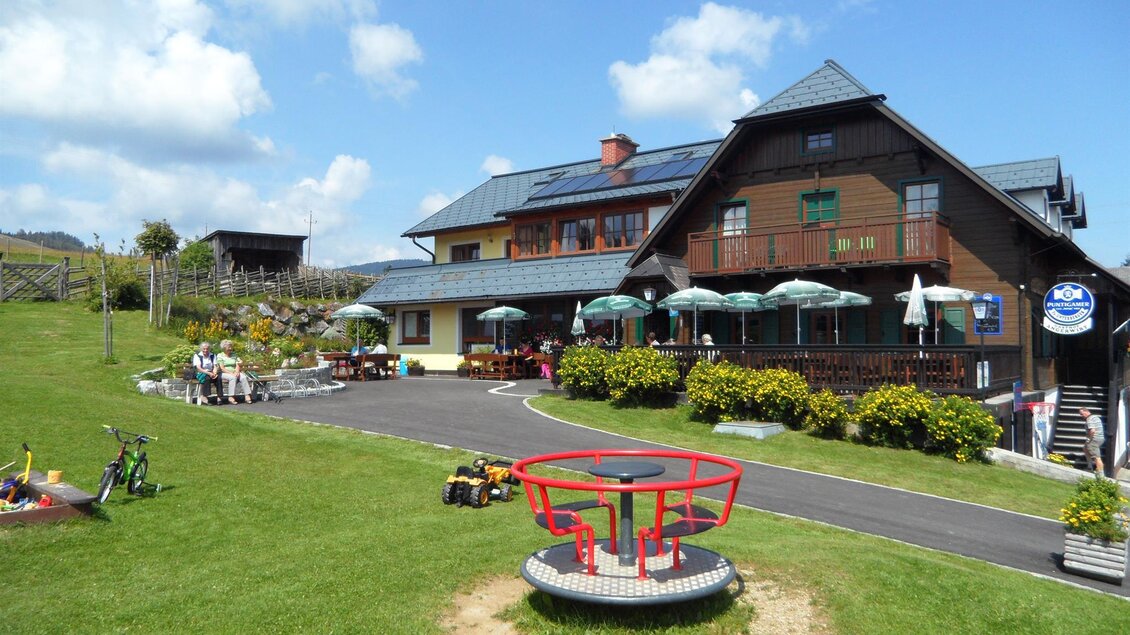 Ein gemütliches Gasthaus mit Terrasse und Sonnenschirmen umgeben von einem grünen Garten. Im Vordergrund gibt es einen Spielplatz mit einer Drehscheibe. | © Almgasthof Angerwirt