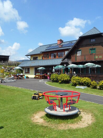 A cozy inn with a terrace and sun umbrellas surrounded by a green garden. In the foreground, there is a playground with a merry-go-round. | © Almgasthof Angerwirt
