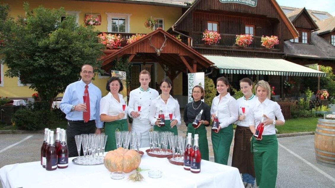 Eine Gruppe von Mitarbeitern steht vor einem Gasthaus und hält Getränke in den Händen. Im Hintergrund sieht man das charmante Gebäude mit blühenden Fenstern. | © Wassermann-Wirt