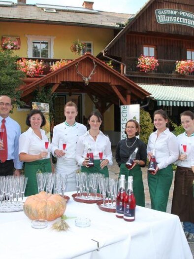 A group of employees is standing in front of an inn, holding drinks in their hands. In the background, you can see the charming building with blooming windows. | © Wassermann-Wirt