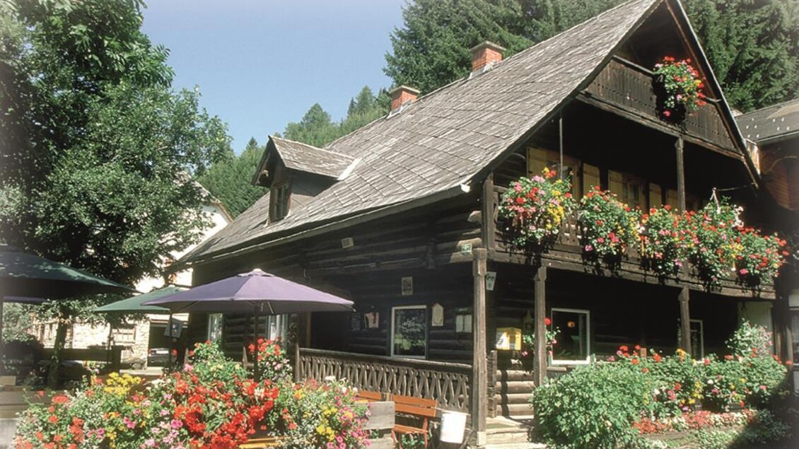 Ein traditionelles Holzhaus umgeben von bunten Blumen und Pflanzen. Im Hintergrund sind hohe Bäume zu sehen, die eine idyllische Atmosphäre schaffen. | © Gasthof Beren "Jägerheim"