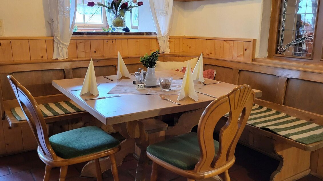 Ein traditioneller Essbereich mit einem Holztisch und Stühlen. Auf dem Tisch sind Servietten und eine kleine Blumenvase dekoriert. | © Petra Kirchschlager