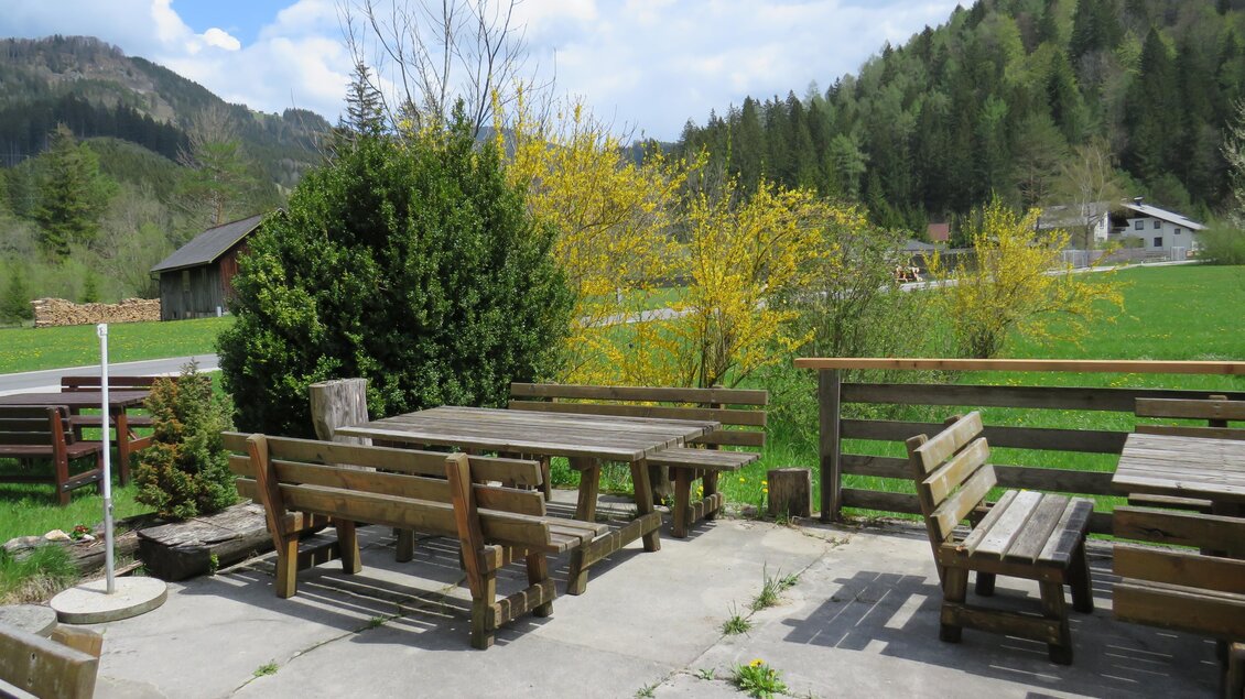 Ein einladender Außenbereich mit Holzmöbeln und einer schönen Aussicht auf die grünen Hügel. Der Himmel ist leicht bewölkt und es blühen gelbe Sträucher im Hintergrund. | © Fam. Digruber