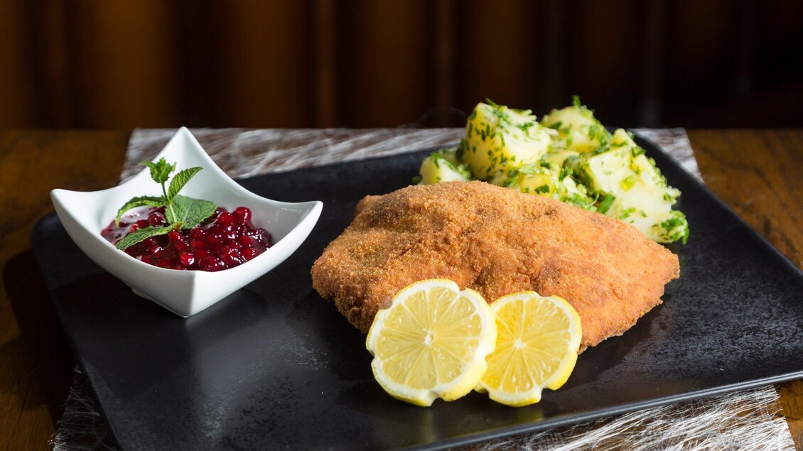 Ein saftiges Schnitzel mit Zitronenscheiben und Beilagen aus Kartoffeln und Preiselbeermarmelade. Das Gericht ist auf einem schwarzen Teller angerichtet. | © TV Region Graz - Rene Vidalli