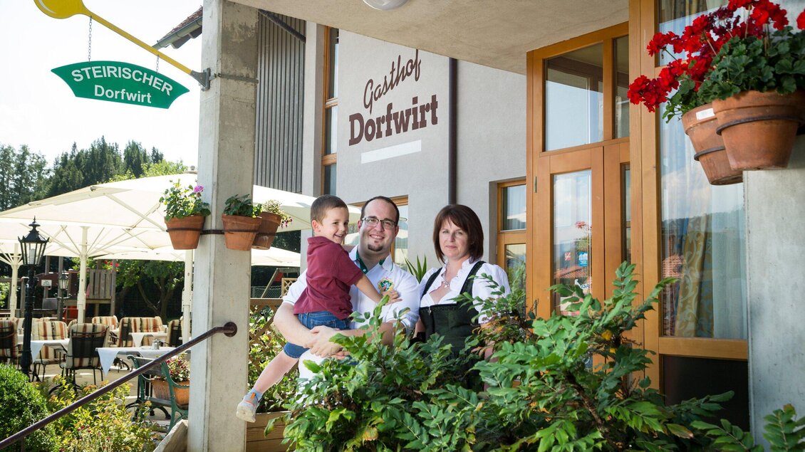 Eine Familie steht vor dem Restaurant "Gasthof Dorfwirt". Im Hintergrund sind Tische und Stühle im Freien sowie Blumen zu sehen. | © TV Region Graz - René Vidalli