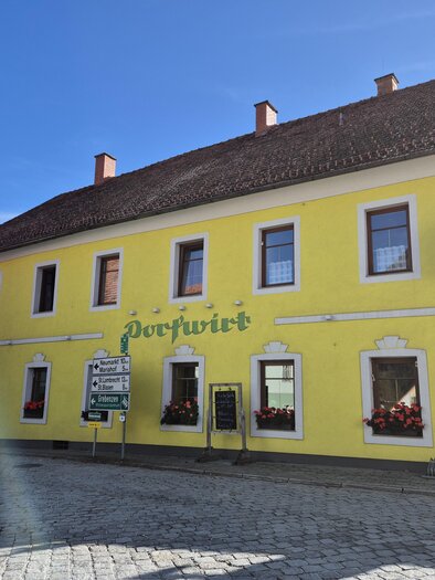 A yellow building with several windows and an awning. In front of the building, tables and chairs are set up outdoors. | © Tourismusverband Murau