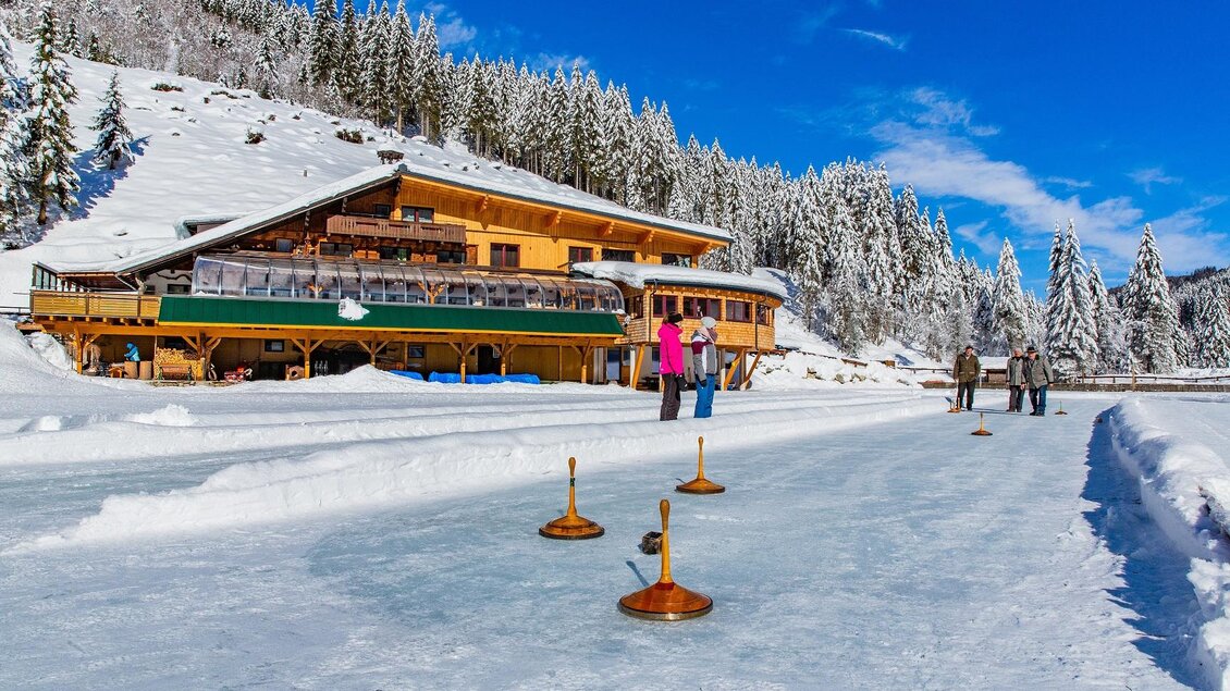 Eine malerische Winterlandschaft mit einer Holzhütte im Hintergrund. Auf dem gefrorenen See spielen Menschen mit Eisstöcken. | © TVB Haus-Aich-Gössenberg@René Eduard Perhab