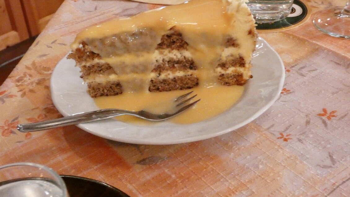 Ein Stück Kuchen auf einem Teller, überzogen mit einer cremigen Soße. Der Tisch ist mit einer blumigen Tischdecke gedeckt. | © Gasthof Franzbauer