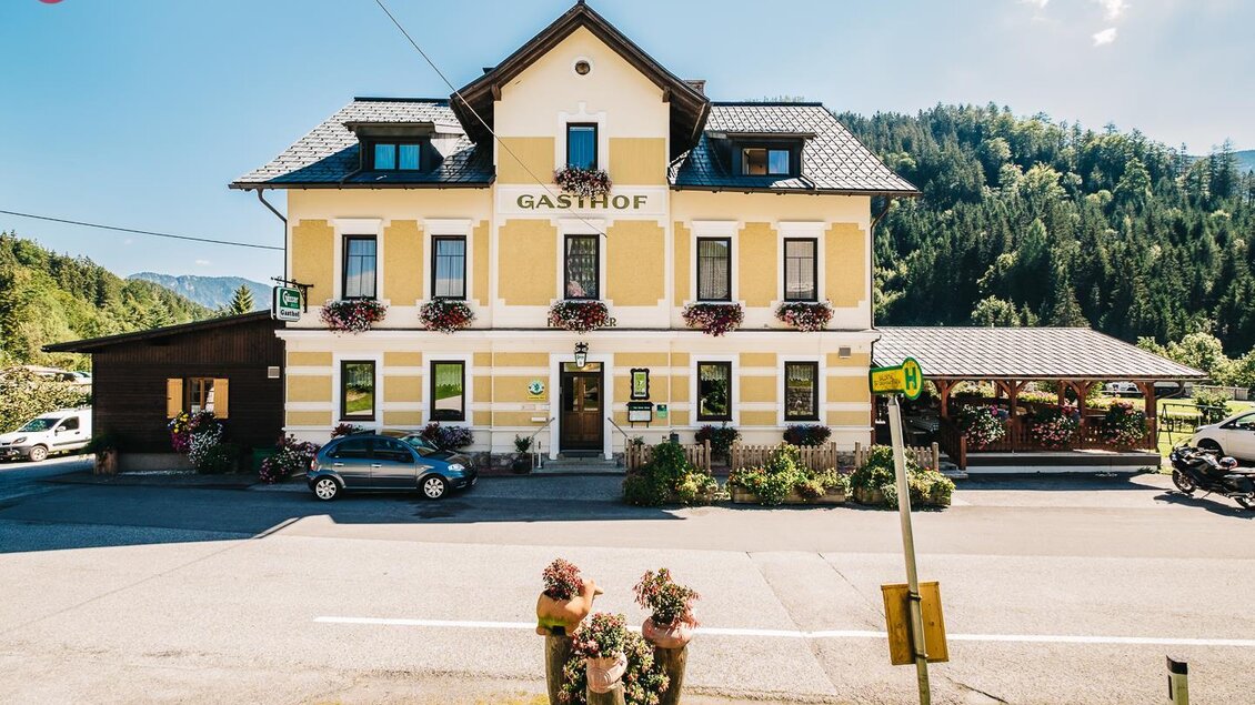 Ein einladender Gasthof mit gelber Fassade und Blumenkästen. Umgeben von grünen Bergen und einer ruhigen Straße. | © Rudy Dellinger